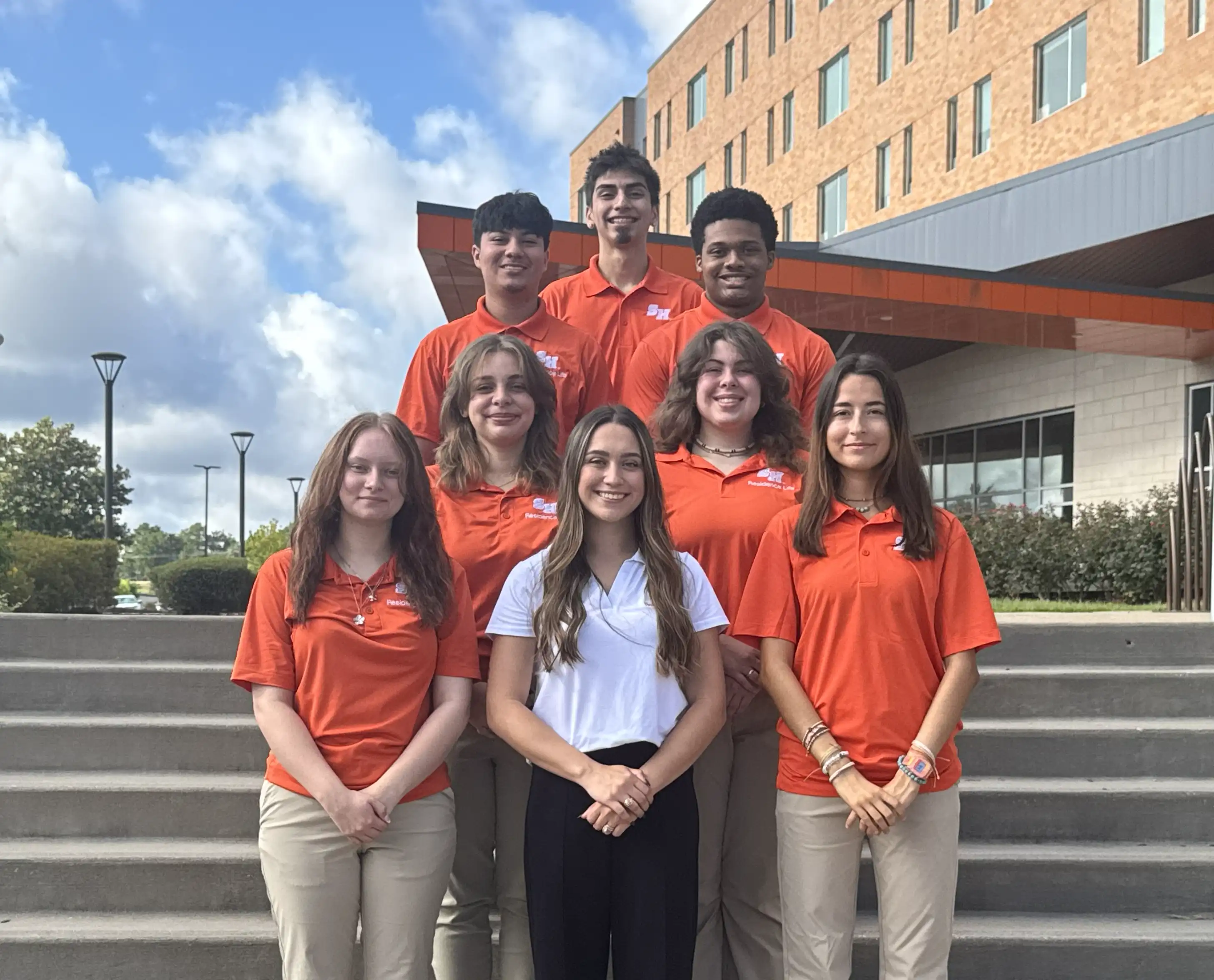 Group photo of Sam Houston State University Residence Life hall staff standing on outdoor steps in front of a residence hall. Most of the staff are wearing matching orange SHSU Residence Life shirts, while one person in the front row is wearing a white floral dress. The group is smiling and posing together for the photo.