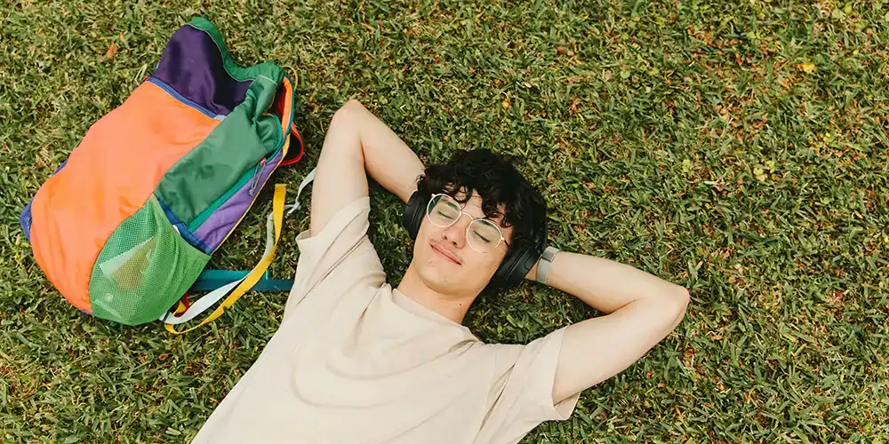 Student lying on grass with eyes closed, wearing headphones and a beige shirt, relaxing next to a colorful backpack.
