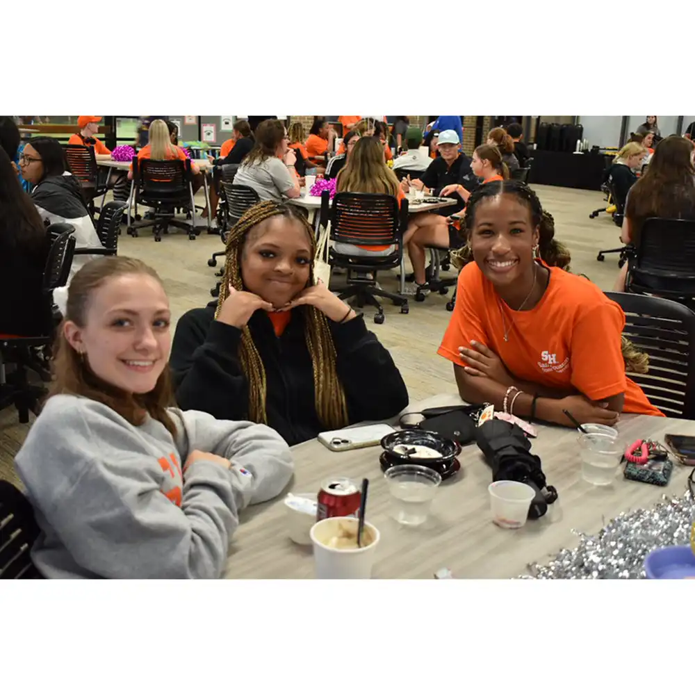 Three SHSU students sit together at a table smiling during a Residence Life event. The room is filled with other students socializing and enjoying snacks.