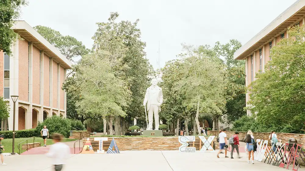 A bright, outdoor scene from the sidelines of the Frank Parker Plaza. A statue of Sam Houston himself stands in the middle of the courtyard, surrounded by lush green trees. Various fraternity and sorority letters and signs are displayed. Students walk and gather in small groups, some heading towards buildings, while others pass by.