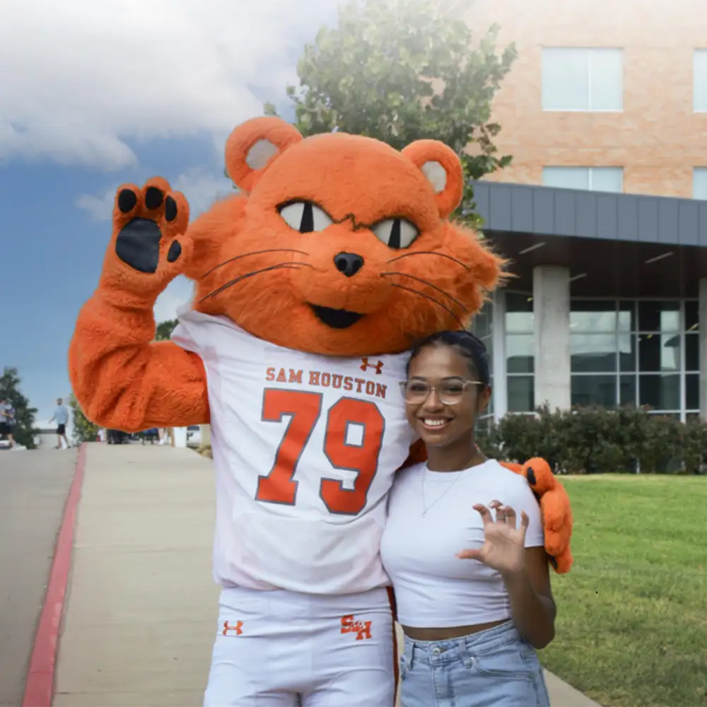 Sammy the Sam Houston State University mascot wearing a white football uniform with the number 79 and “Sam Houston” printed on the chest. Sammy is standing outdoors in front of Piney Woods Hall, raising one paw in a "Eat 'Em Up" stance and placing the other arm around a person. The person is wearing a white top and light blue jeans and is making a claw gesture with one hand. The background shows a sidewalk, greenery, and part of Lone Star Hall.