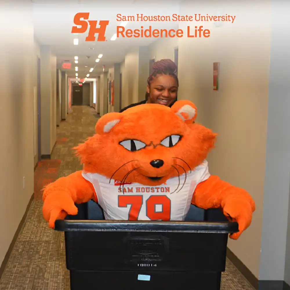 Exterior view of Piney Woods Hall entrance at Sam Houston State University. The building features large glass windows and an orange accent above the doorway with the words “Piney Woods Hall” displayed prominently. Several people are moving items inside using blue rolling carts and carrying bags. The scene suggests a move-in day.