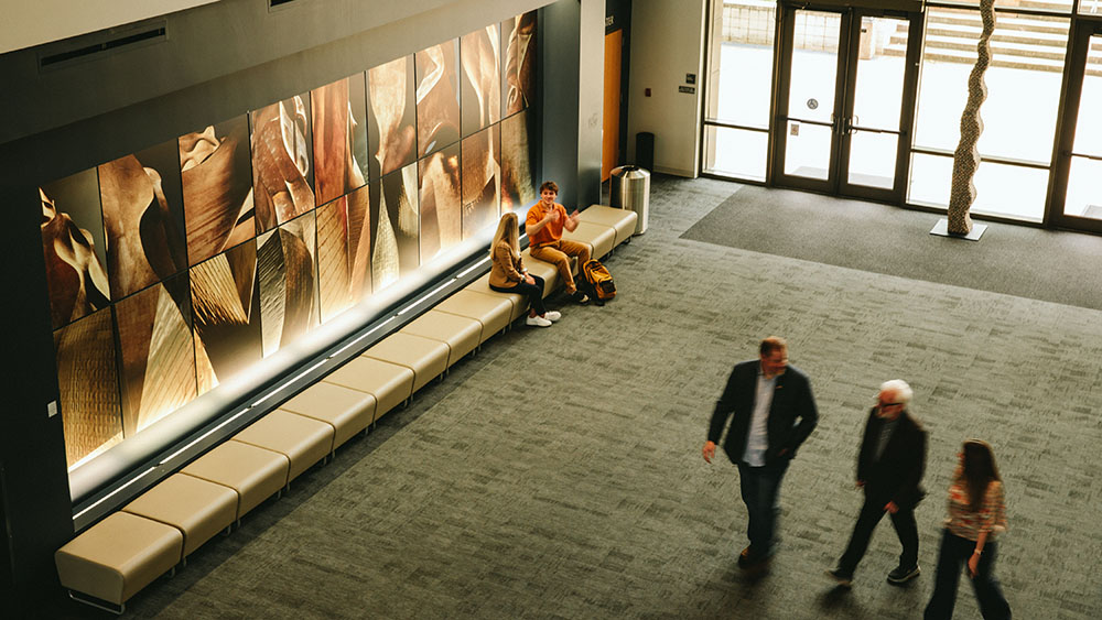 Students sit on a bench beneath a large abstract wall display in a modern lobby while others walk across the open floor near glass doors.