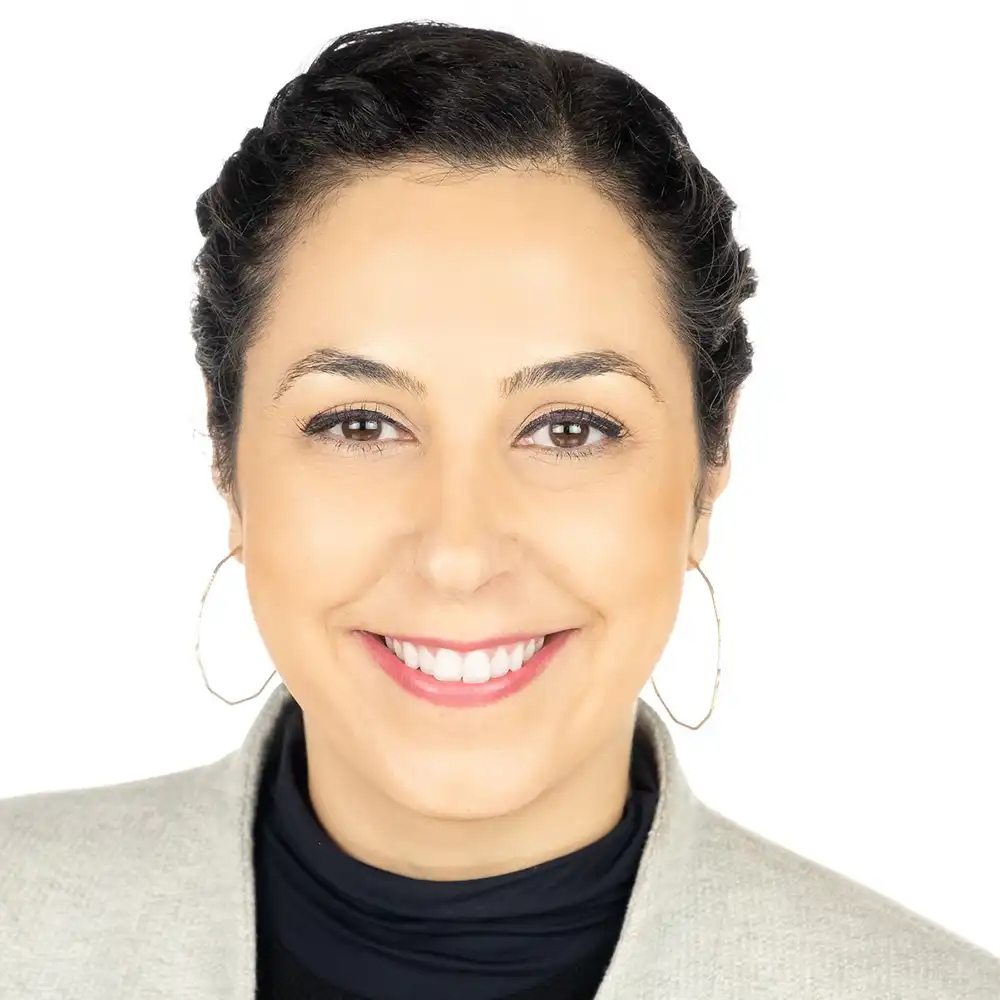 Woman smiling outdoors, standing with hands on hips, wearing a yellow patterned blouse and black pants, with greenery in the background.