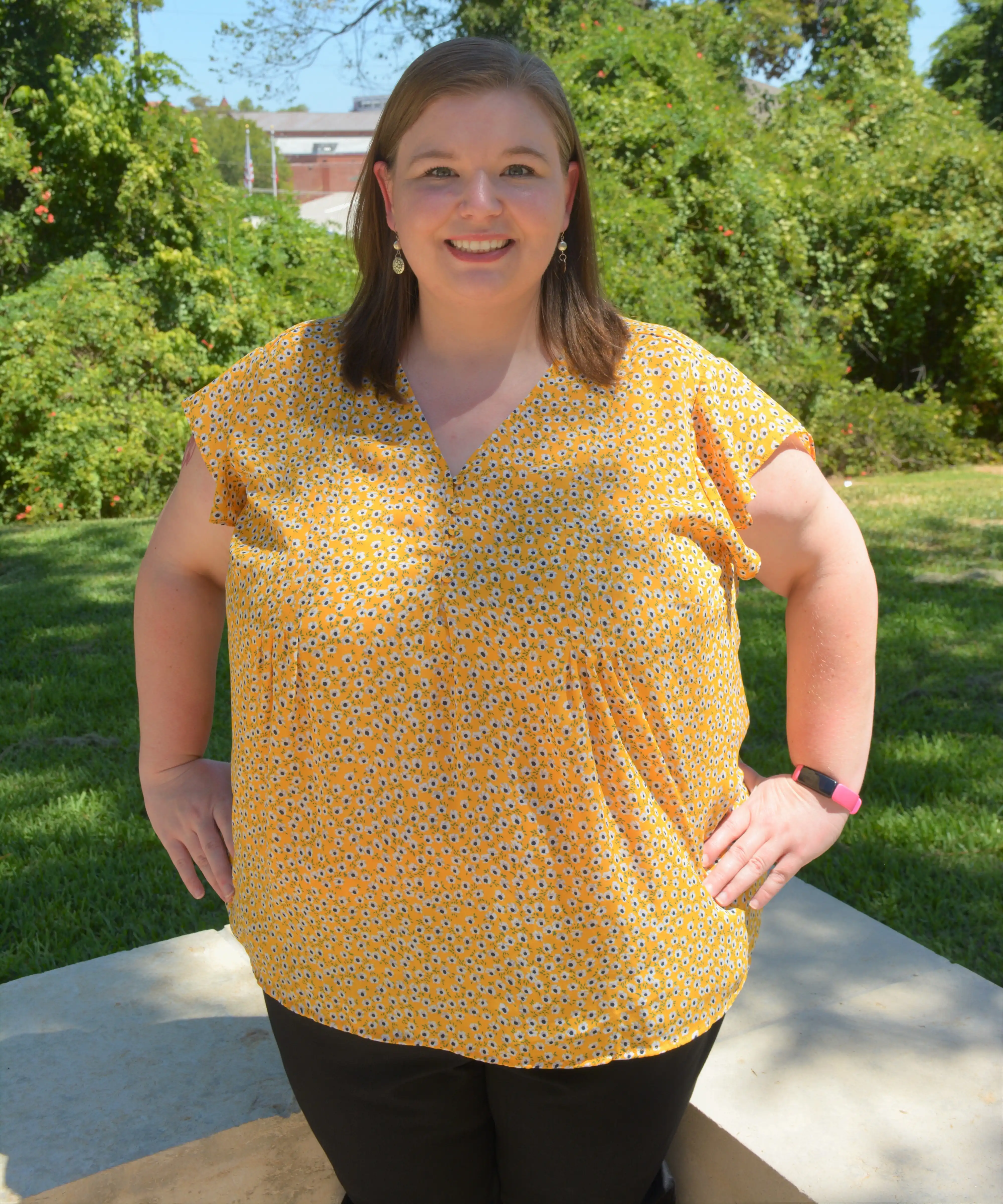 Woman smiling outdoors, standing with hands on hips, wearing a yellow patterned blouse and black pants, with greenery in the background.