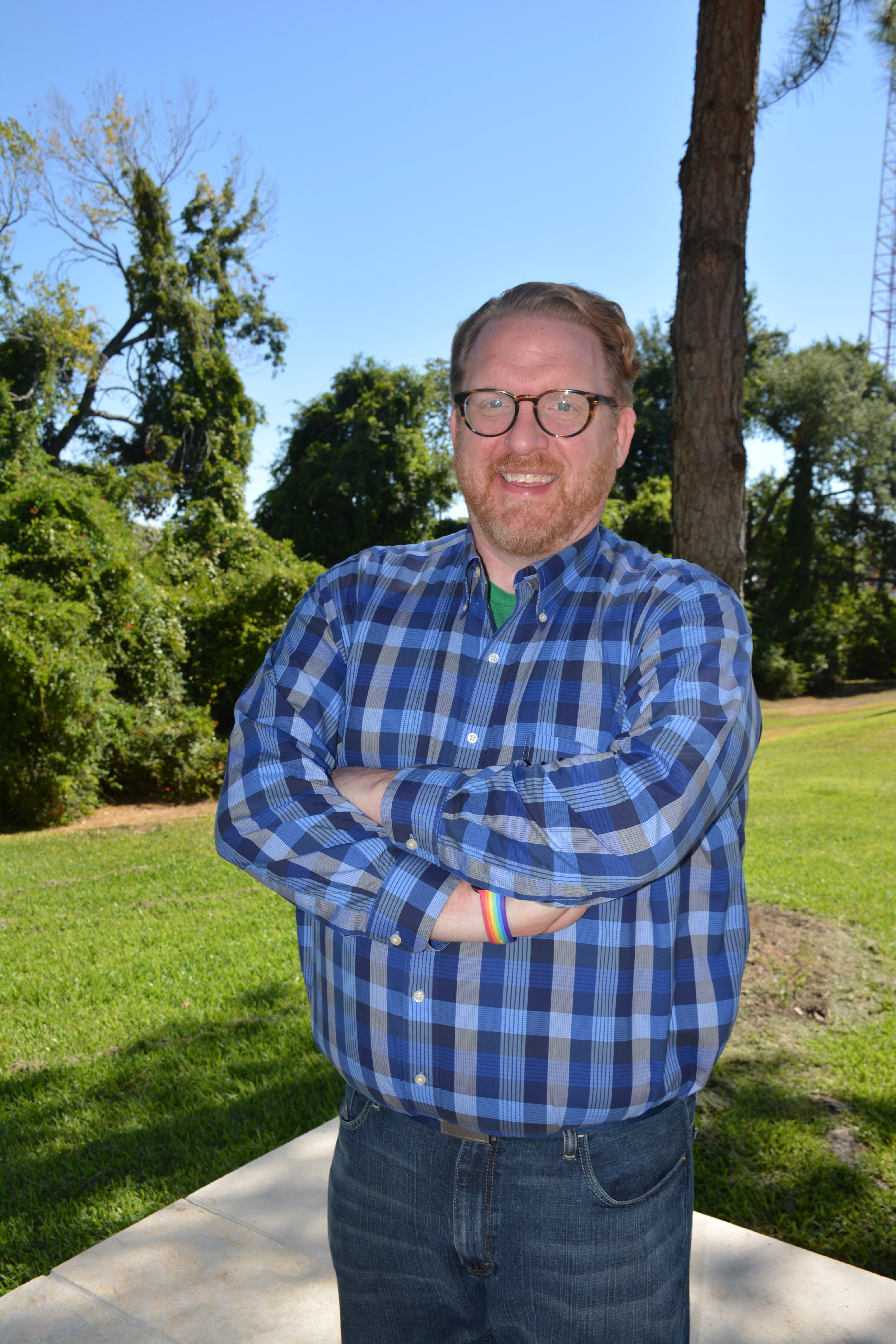 A smiling man with glasses and a beard stands outdoors with arms crossed, wearing a blue plaid shirt and a rainbow wristband, surrounded by trees and blue sky.