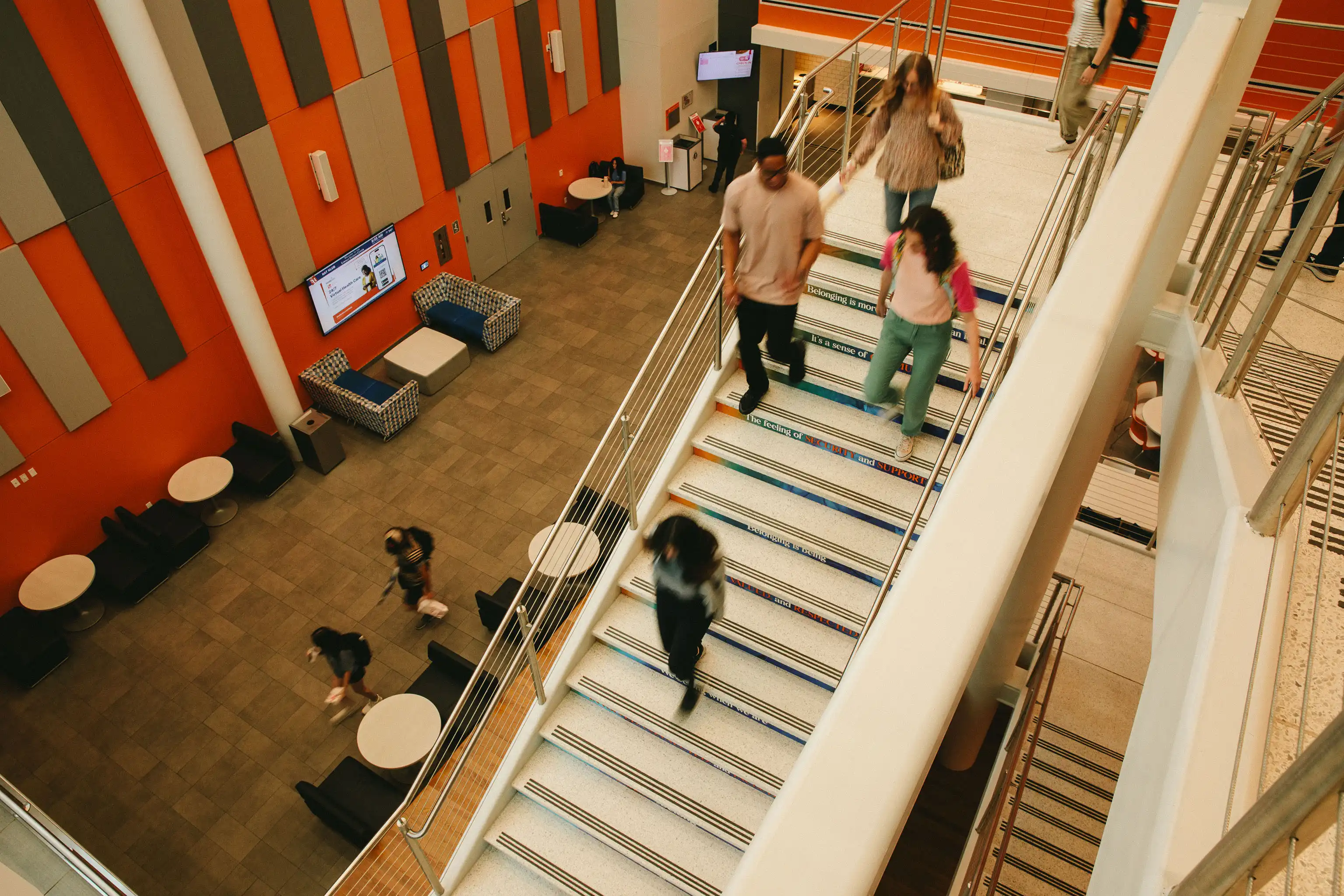 Students walk up and down a brightly lit staircase inside the Lowman Student Center with orange and gray wall panels, lounge seating, and round tables below.