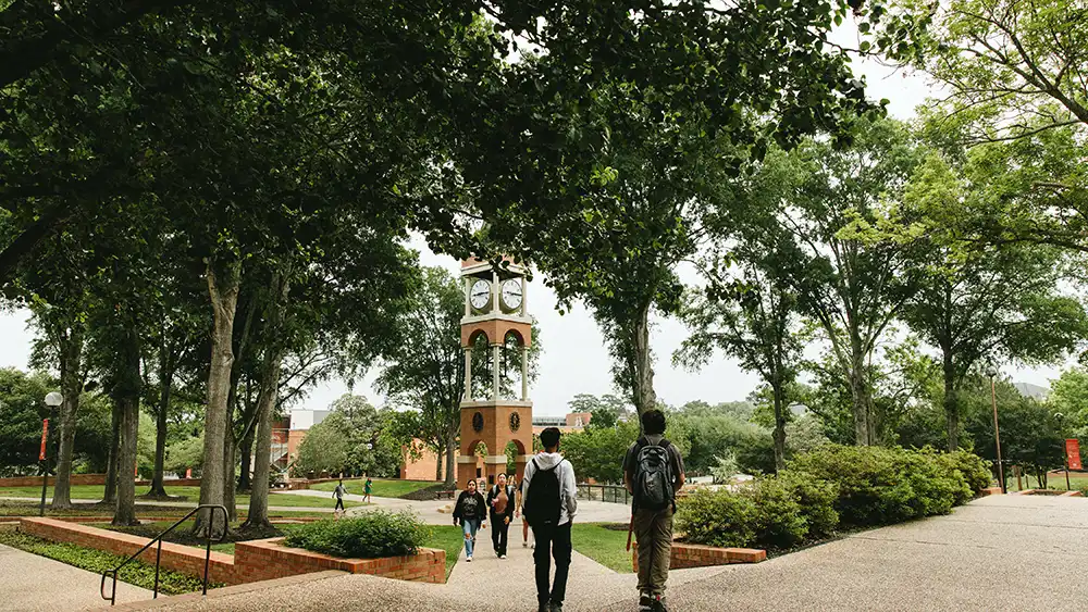 Students walk along a tree-lined pathway on the Sam Houston State University campus, with the iconic clock tower visible in the background. Brick planters, green landscaping, and shaded sidewalks frame the scene, creating a welcoming campus atmosphere on a bright day.