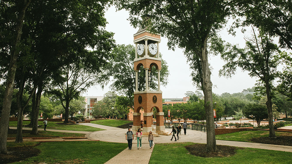 Students walk past the brick bell tower on the Sam Houston State University campus, surrounded by green trees and paved walkways.
