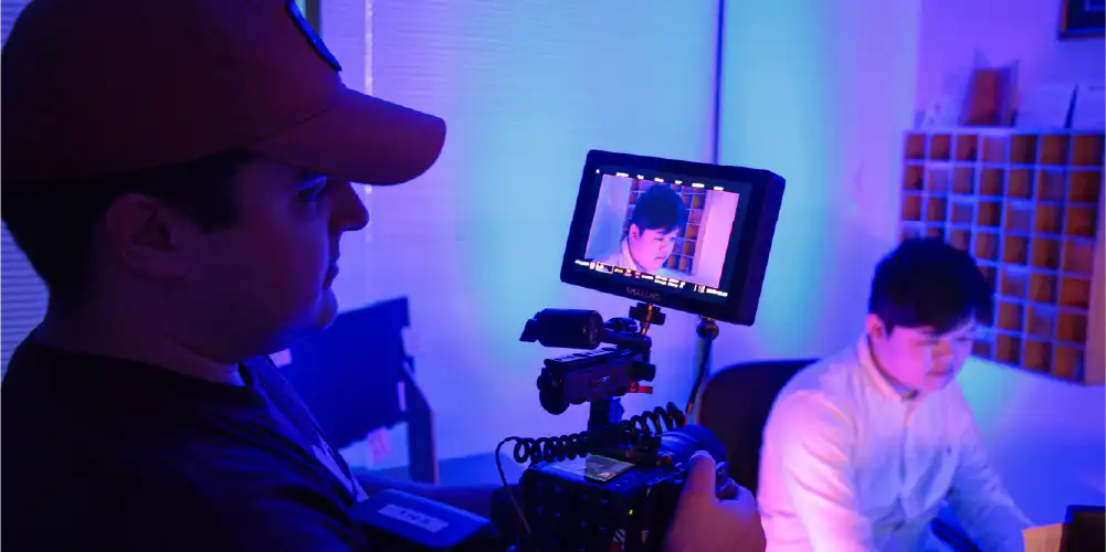 Student holding RED camera on set while filming a in a blue light with an actor in the background and on the camera monitor.