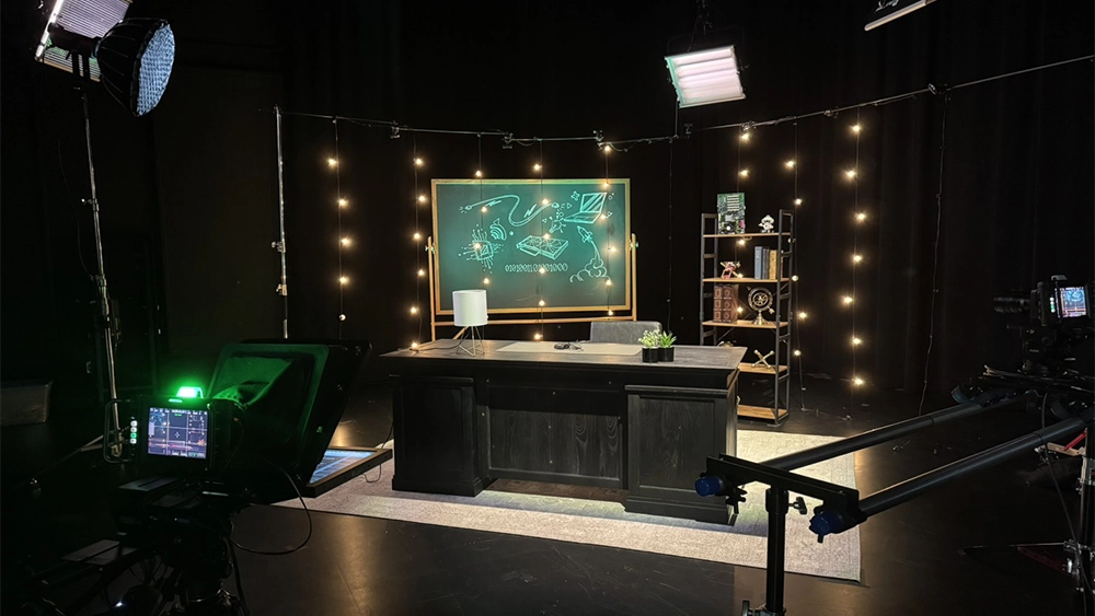 Dark film studio with a camera in the foreground and a desk on a carpet set up in the middle of the room. Lights are around the set lit up.