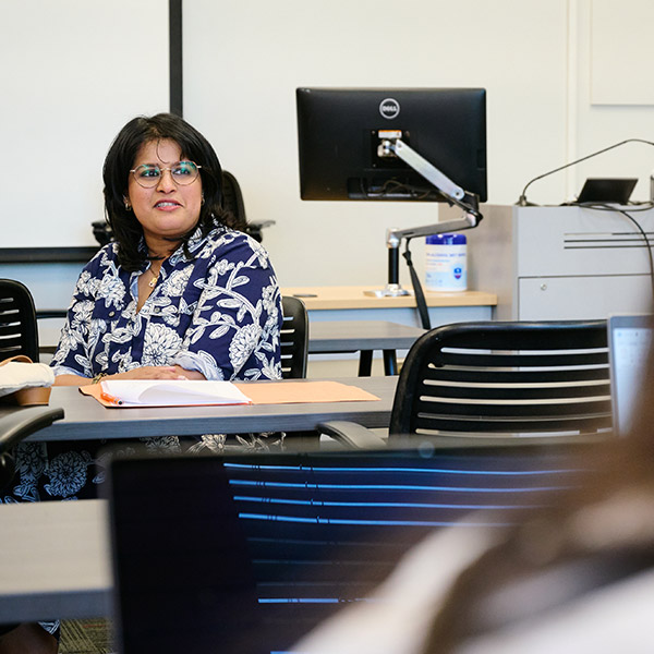 A woman with dark hair and glasses, wearing a blue and white floral patterned blouse, sits at a desk in a classroom or office setting. She is looking toward someone off-camera with a slight smile.