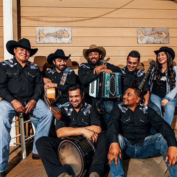 A group portrait of seven members band. They are dressed in matching black western shirts with decorative silver patterns on the shoulders, jeans, and cowboy hats. Several members hold instruments, including an accordion and a snare drum. They are smiling and laughing together against a tan wooden wall decorated with two metal fish skeleton art pieces.