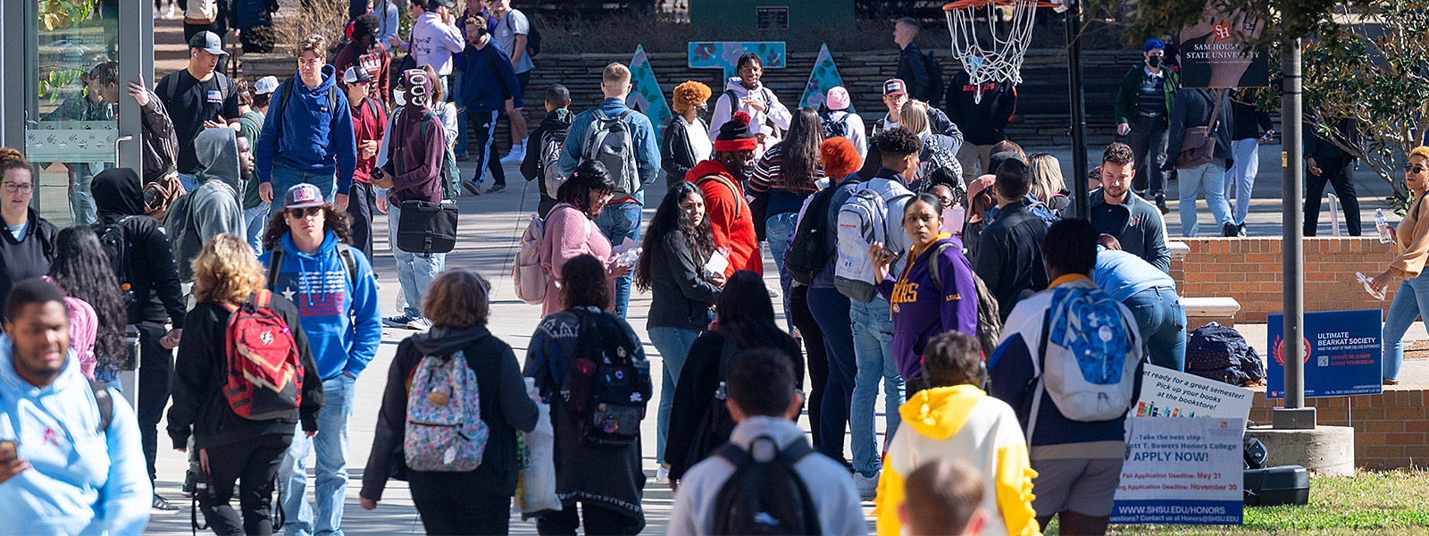 A crowded outdoor campus plaza at Sam Houston State University. Students are walking in various directions. In the center stands a large white statue of Sam Houston. To the right, a basketball is captured mid-air near a hoop. Trees and campus buildings are visible in the background under bright sunlight.
