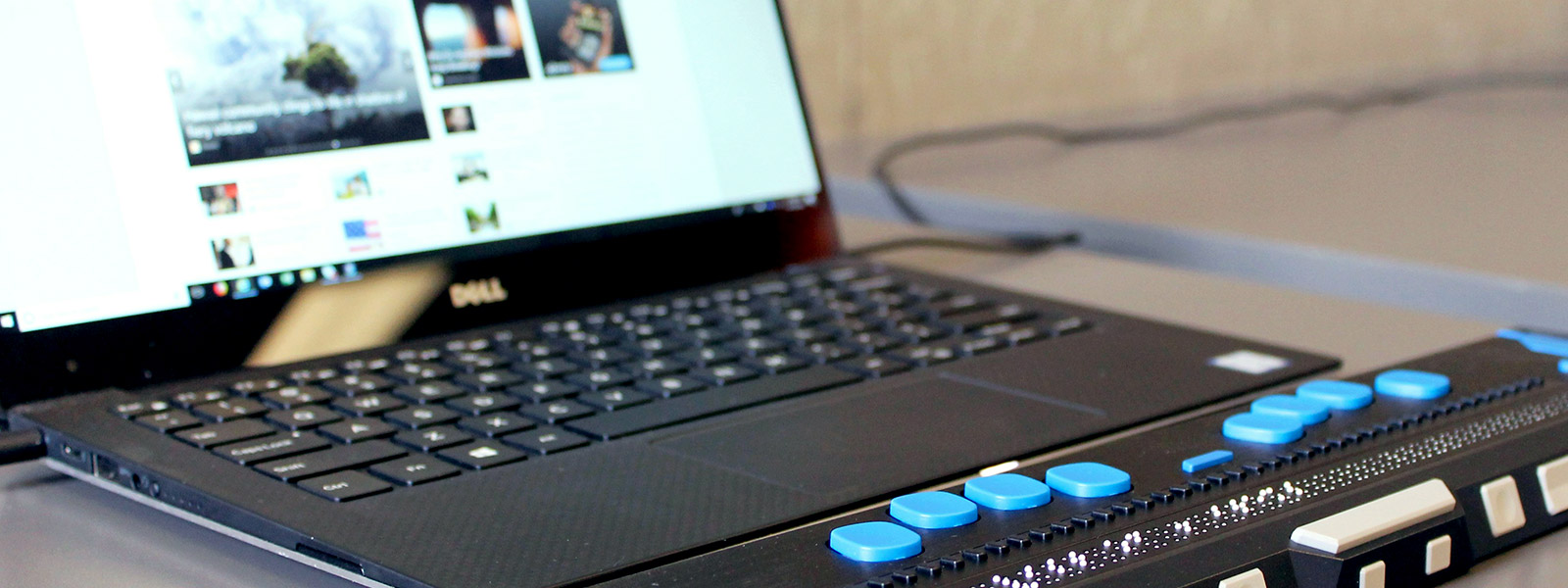A close-up of a laptop connected to a Refreshable Braille Display sitting on a gray desk. The braille display is black with blue buttons and tactile braille pins raised in the center. The laptop screen shows a webpage with various tiles and images.