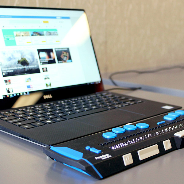 A close-up of a laptop connected to a Refreshable Braille Display sitting on a gray desk. The braille display is black with blue buttons and tactile braille pins raised in the center. The laptop screen shows a webpage with various tiles and images.