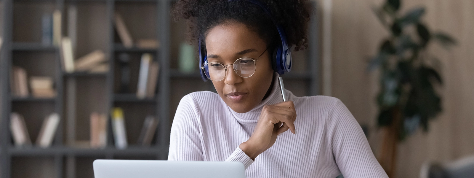 Woman in her 30s listening to course content through headphones and looking at a laptop. She has a pen in her hand.