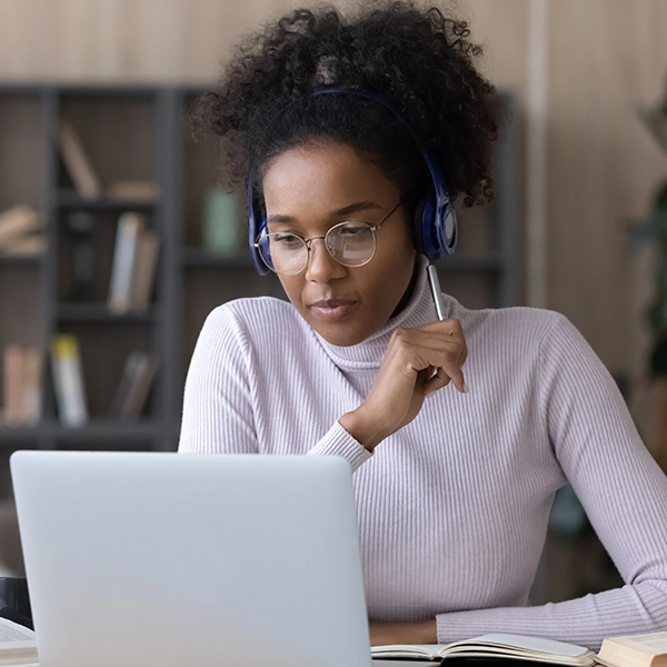 A woman wearing glasses and headphones looking at a laptop while holding a pen