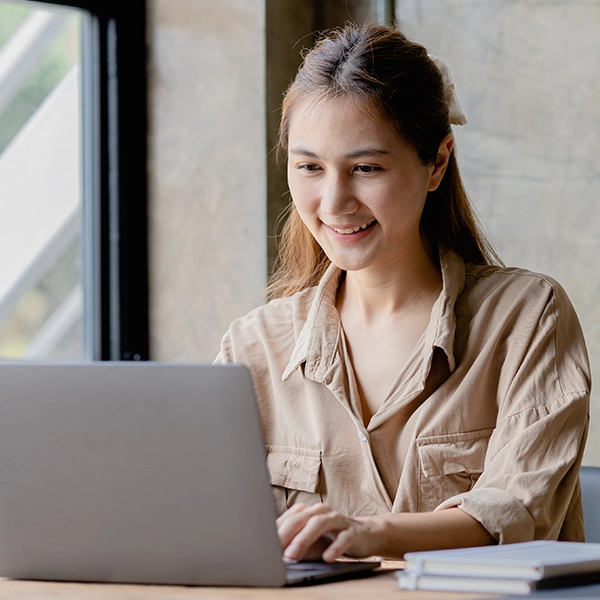 A woman smiling while typing on a laptop at a desk with notebooks