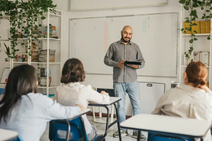 A smiling teacher holds a tablet and stands before a whiteboard in a bright classroom, facing three seated students.
