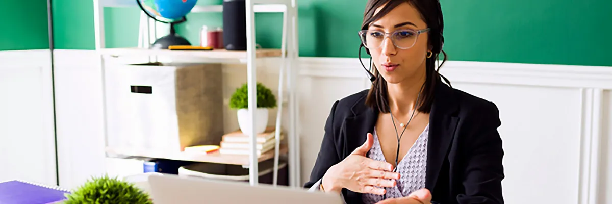 A woman speaks during a video call from a home office, seated behind a laptop with books and office decor in the background.