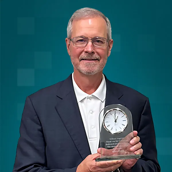 Dr. Bill Angrove smiles holding a glass clock award against a teal background