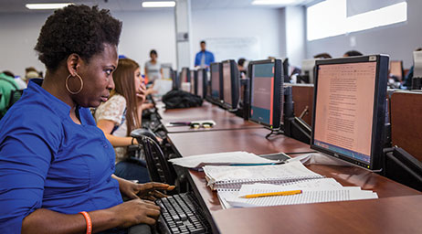 Students working on computers with focus on a woman writing a document on one of the computers.