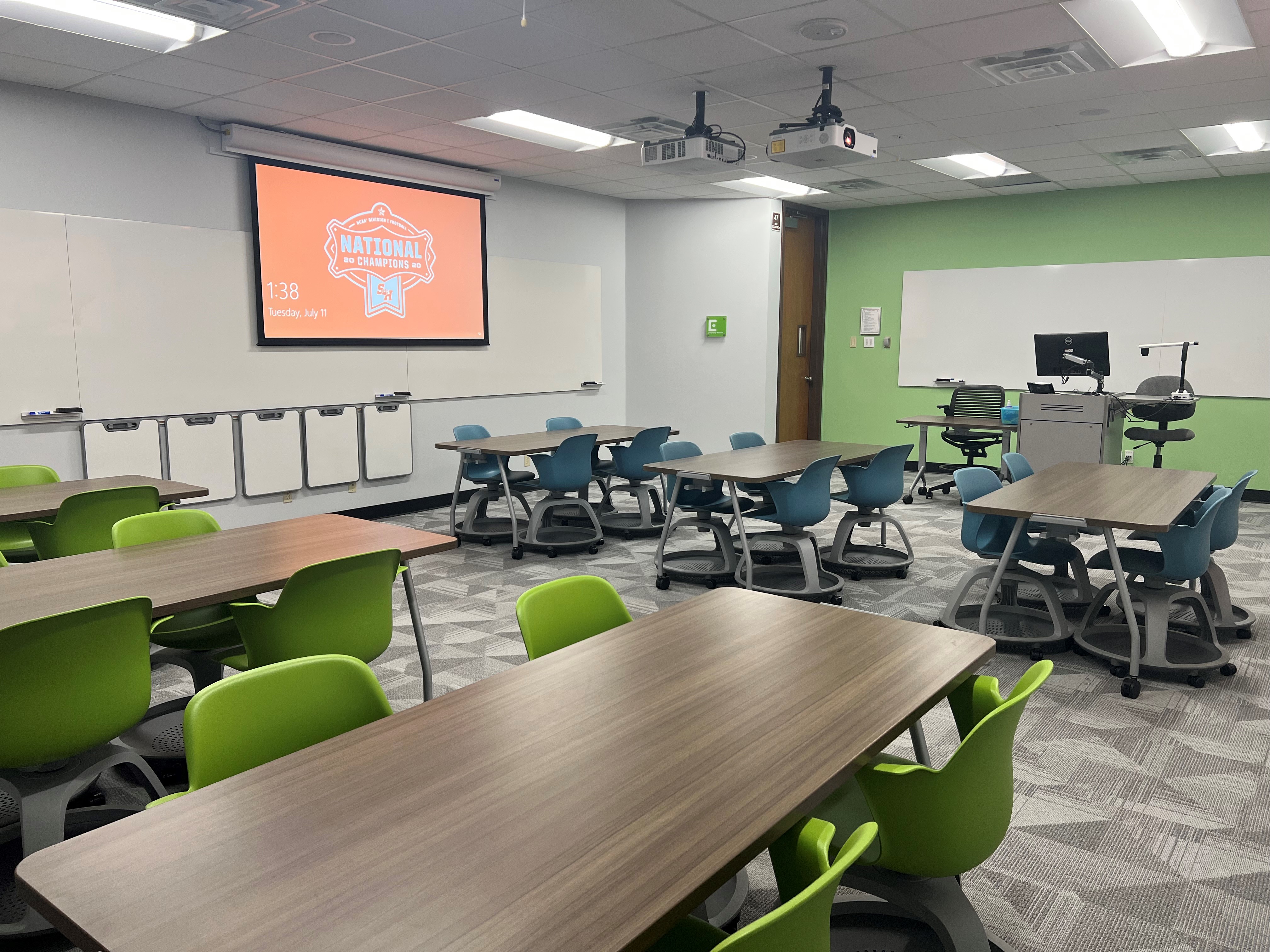LDB 208, tables and chairs in a  classroom with a projector screen in the background.