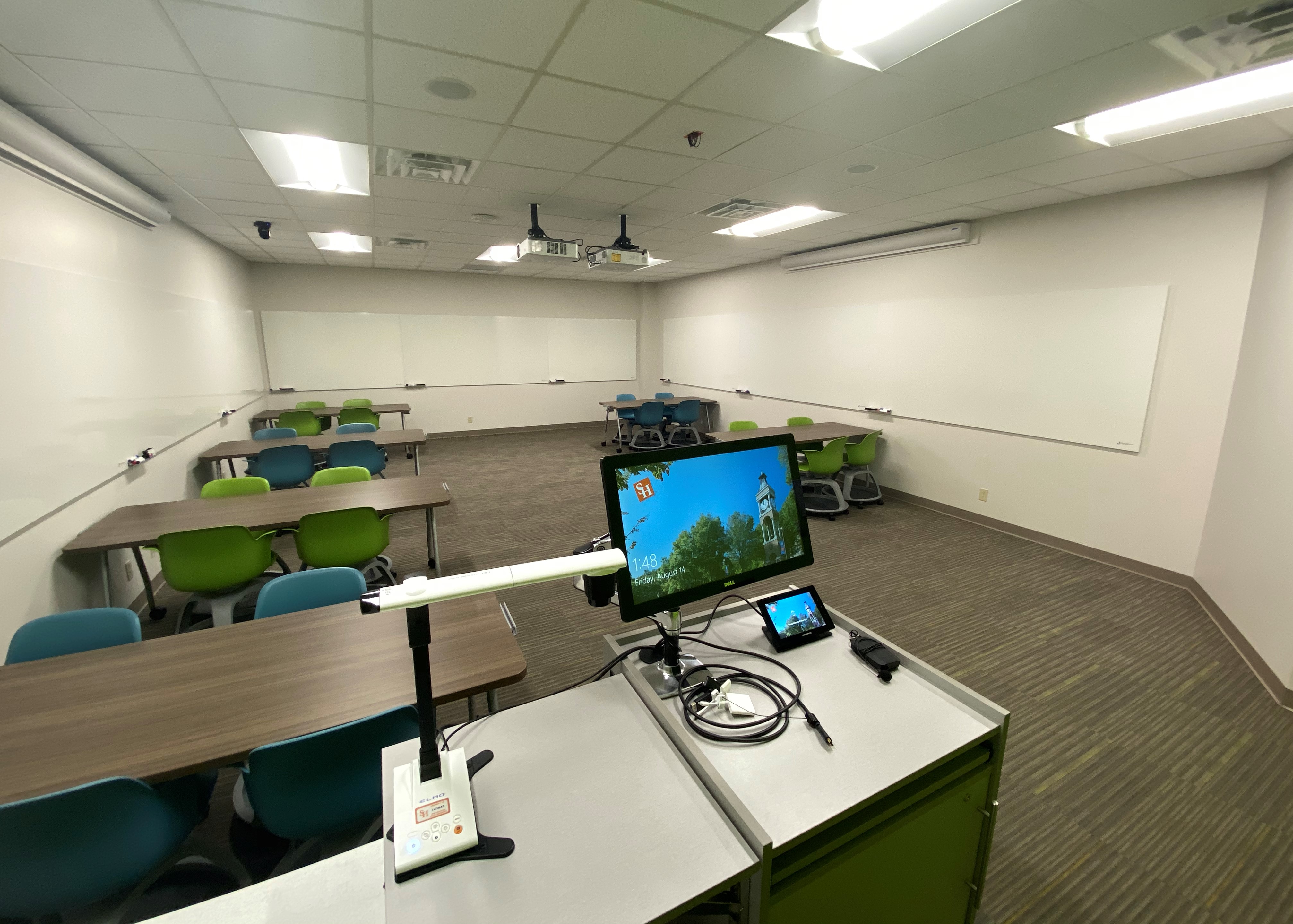 LDB 208, tables and chairs ina large classroom with a projector screen in the background. One wall is bright green and the other is white.