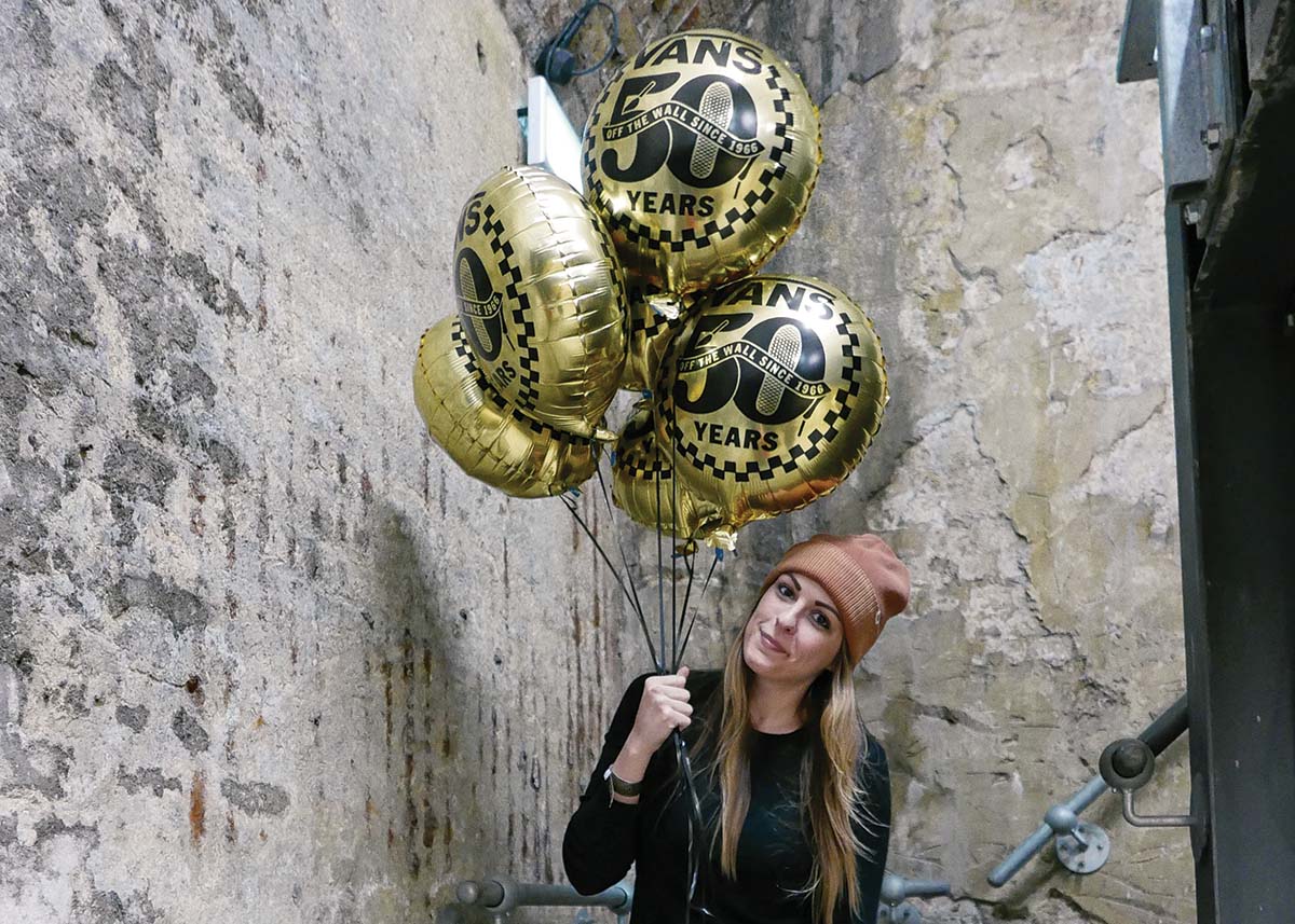 Amanda Silverman stands in a concrete stairwell and holds golden balloons with logos that indicates Vans 50 Year Anniversary.