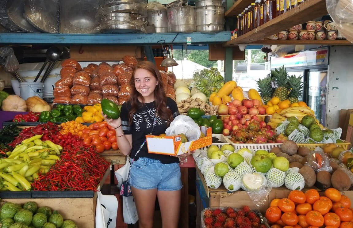 Taylor Pennington poses with a large assortment of brightly colored vegetables in a covered outdoor market.