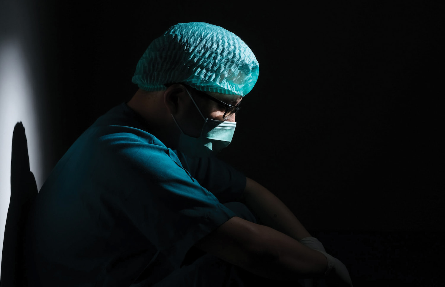 A person wearing surgical scrubs and a mask sits against a wall in a dark, clean room.