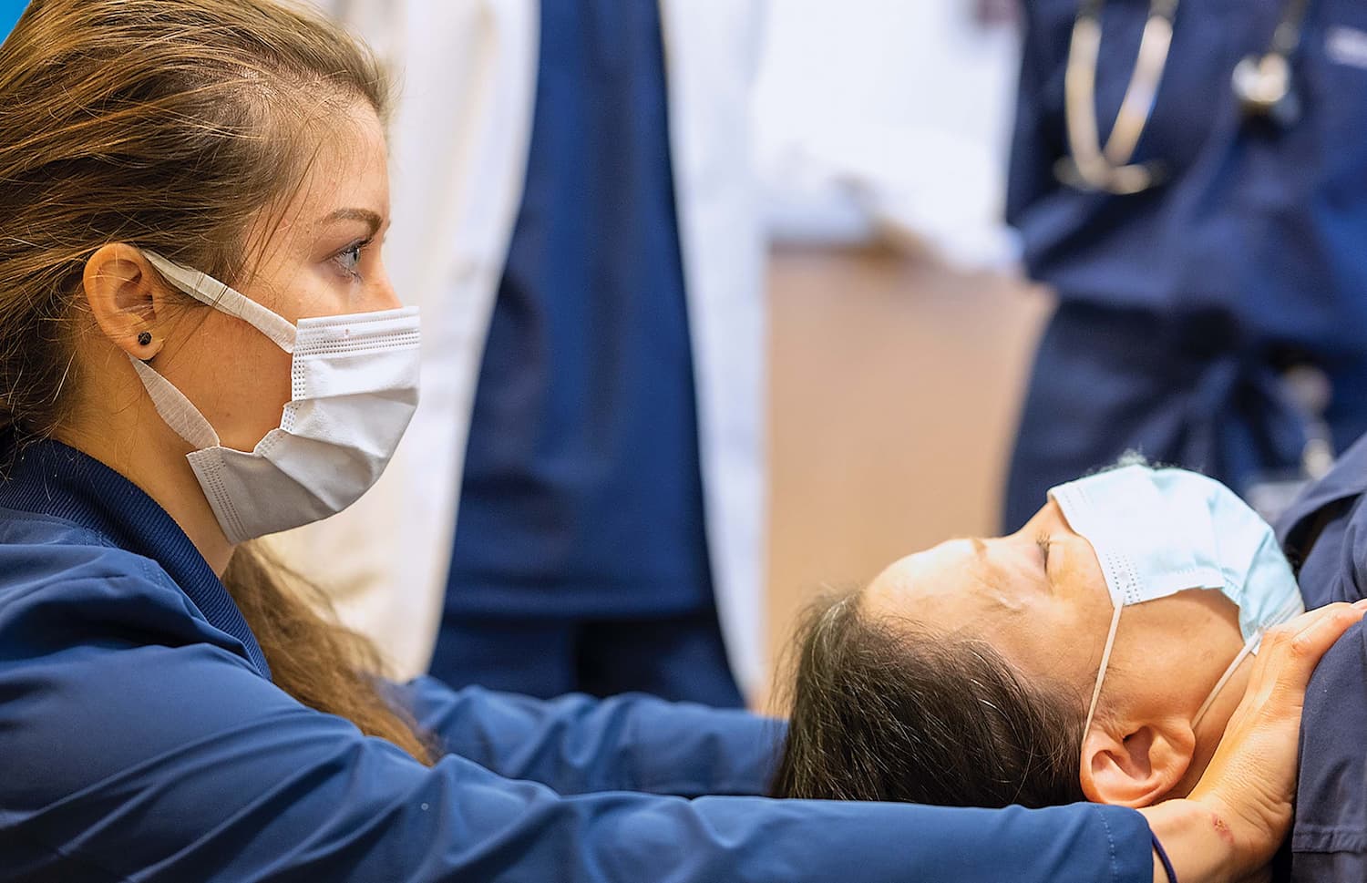 A person in scrubs and wearing a mask massaging the shoulders of another person that is laying down, also wearing a mask.