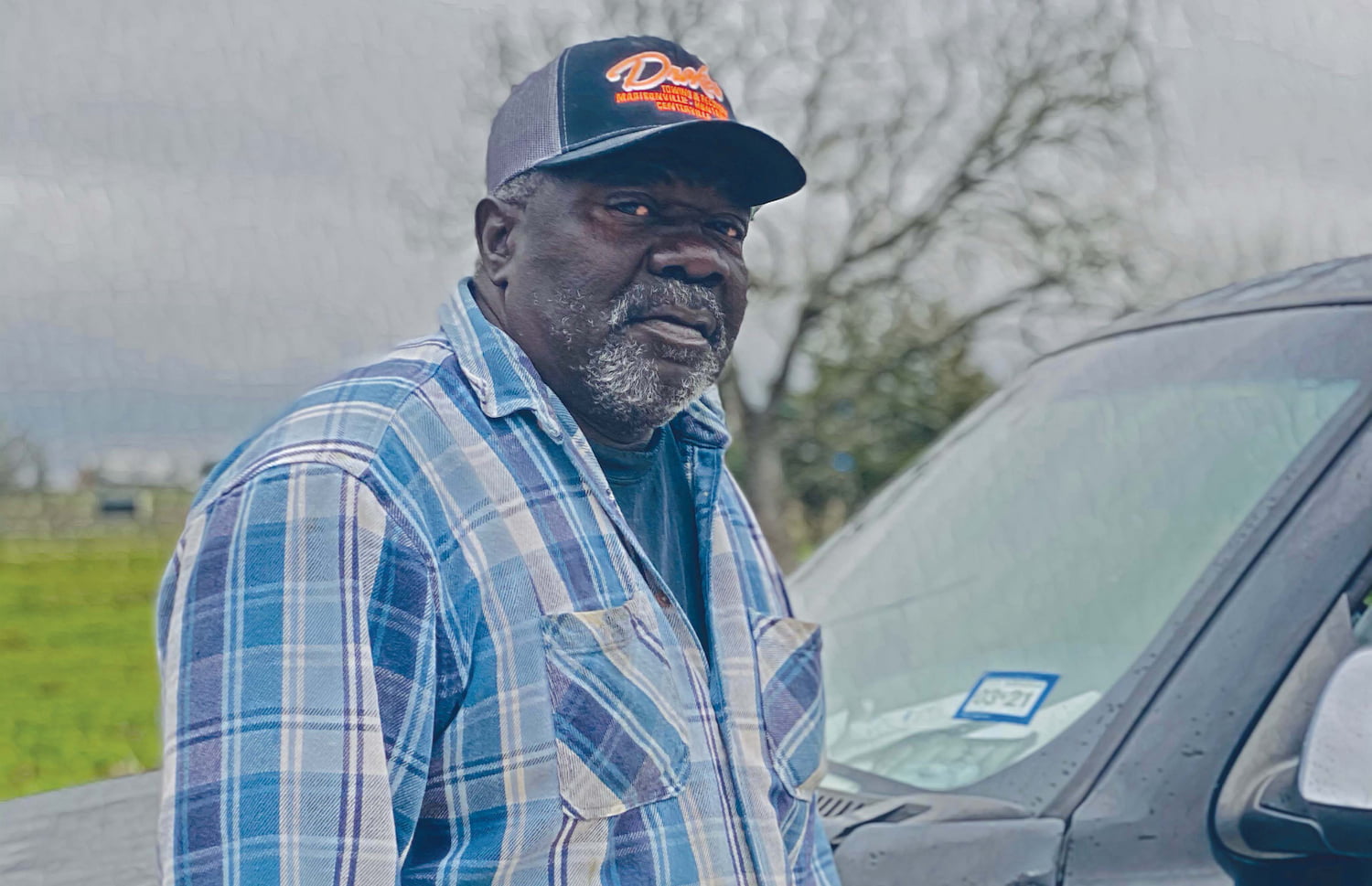 George Dunn stands next to his pickup truck in overcast weather. He wears a baseball cap and a button up shirt.