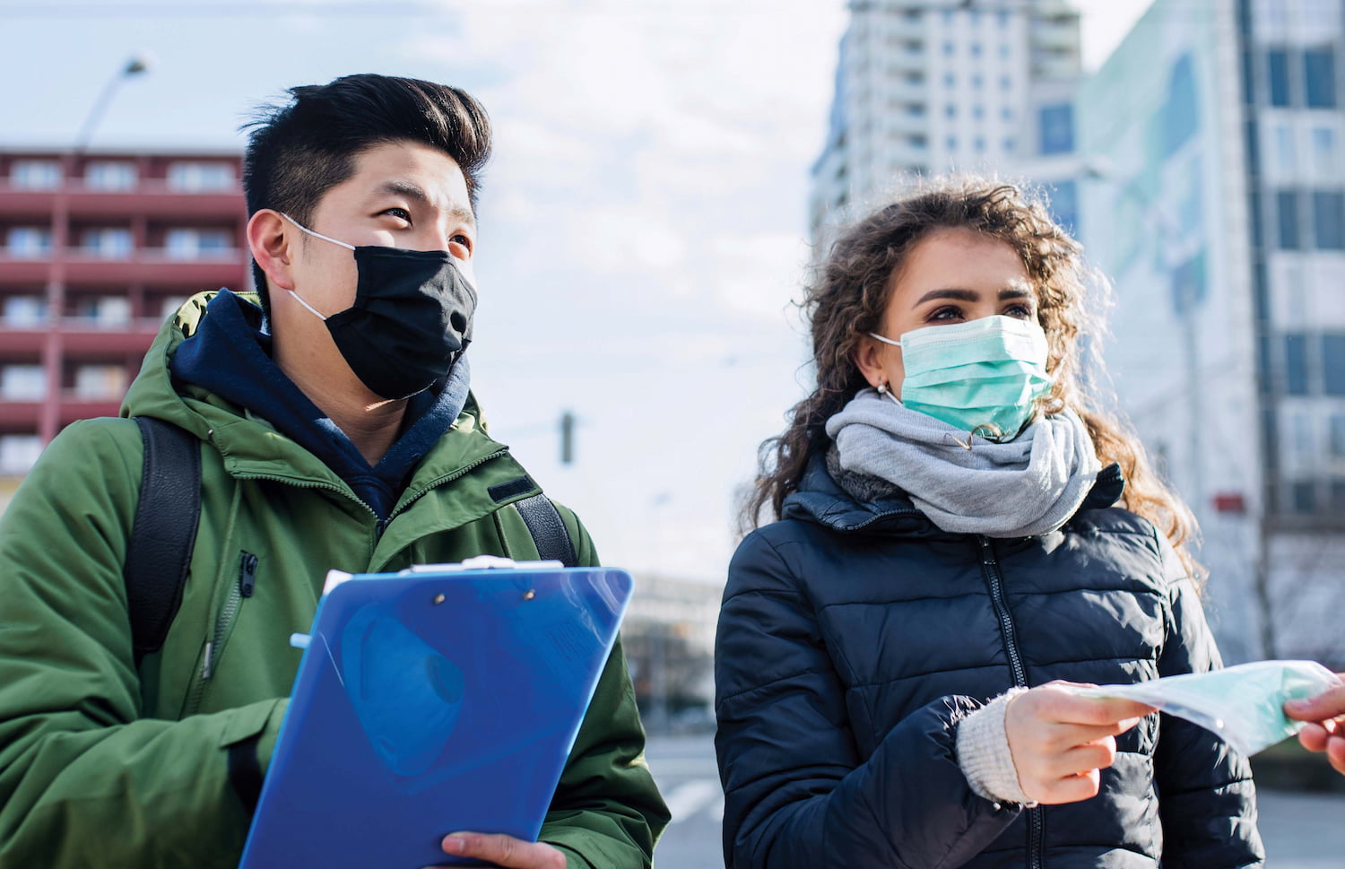 One person holds a clipboard while the other hands out masks outdoors in the city.