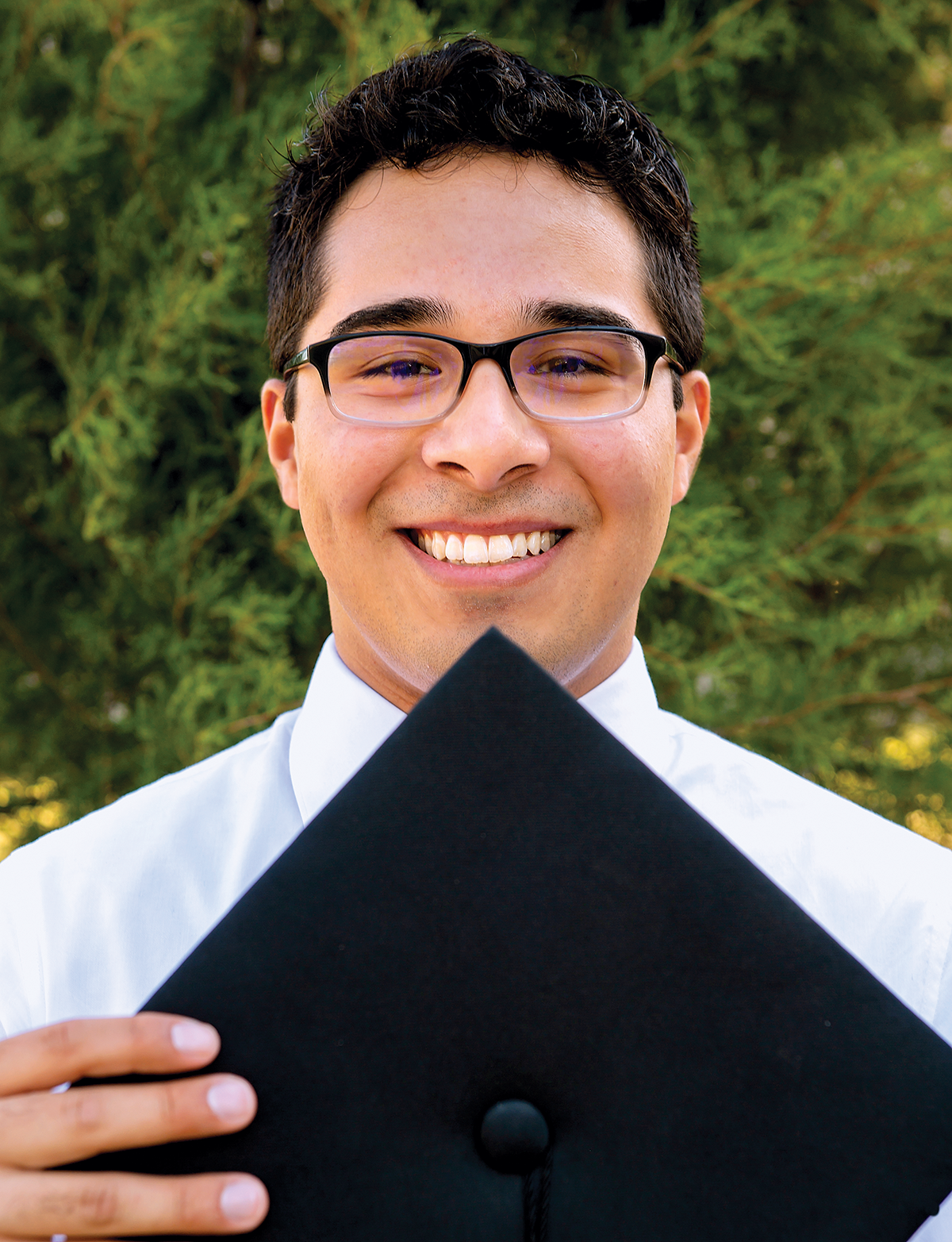 Student, Jesus Martinez, poses with a graduation cap and looks directly at the camera. There is greenery behind him.