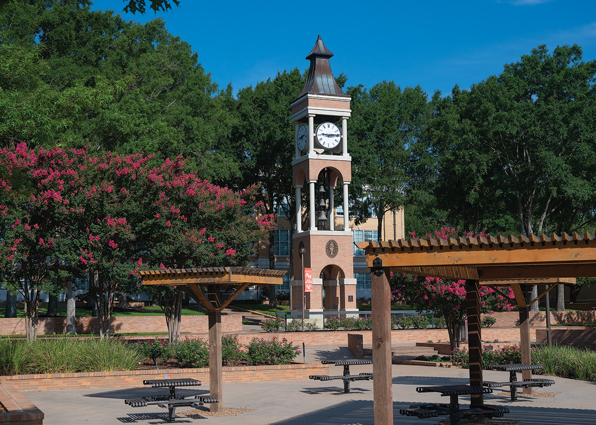 The SHSU Clocktower stands tall against the green trees. Beautifiul pink flowers are on either side of it. In the foreground, benches and wooden structures for shade exist.